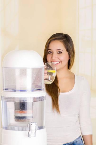 Beautiful woman drinking a glass of water with a filter system of water purifier on a kitchen background - Photo, image