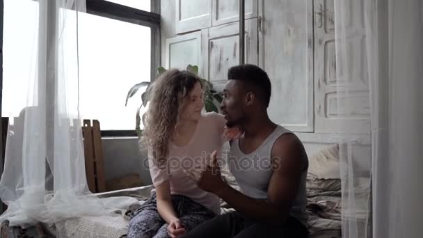 Beautiful multiethnic couple in pajamas sitting on bed and talking. Man and woman have a conversation. Slow motion. - Video