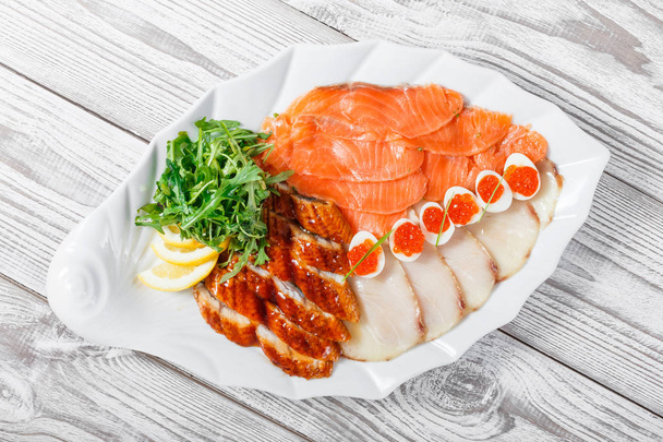 Seafood platter with salmon slice, smoke sturgeon, quail eggs with red caviar, slices fish fillet, decorated with arugula and lemon on wooden background close up. Mediterranean appetizers. Top view - Photo, Image