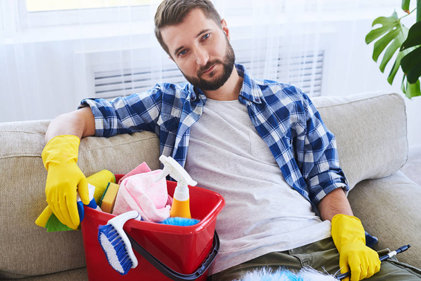 Tired guy sitting on sofa with cleaning set - Photo, Image