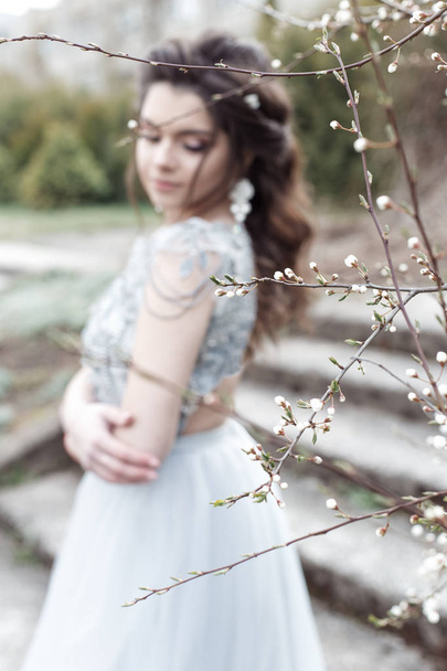 Beautiful young tender gay girl with a beautiful wedding hairstyle with a snow-white smile in a light dress is walking in a spring blooming garden - Foto, Bild