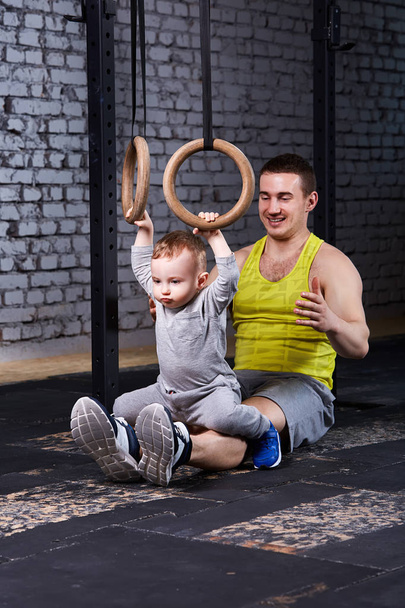 Smiling young father trains the little son with gimnastic rings against brick wall in the gym. - Fotoğraf, Görsel