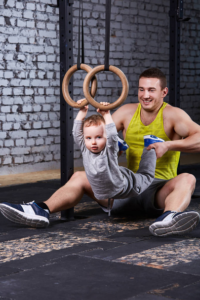 Smiling young father trains the little son with gimnastic rings against brick wall in the gym. - Fotó, kép