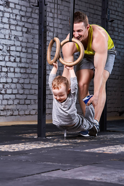 Young sporty father trains the little son with gymnastic rings against brick wall at the cross fit gym. - Fotografie, Obrázek
