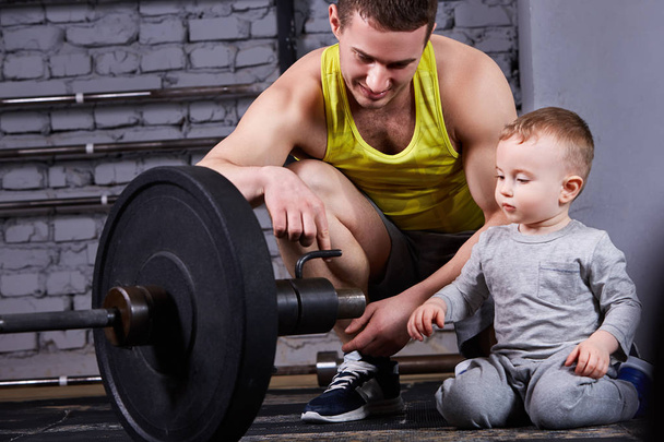 Young smiling father shows little son the dumbbell against brick wall at the cross fit gym. - Φωτογραφία, εικόνα