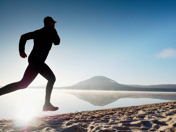 Gezonde levensstijl. Hoog jongeman runner draait op zonsopgang aan zee. Man sprong in zand - Foto, afbeelding