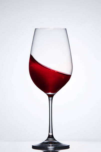 Moving red wine in the elegant wineglass standing against light background with reflection. - Photo, Image