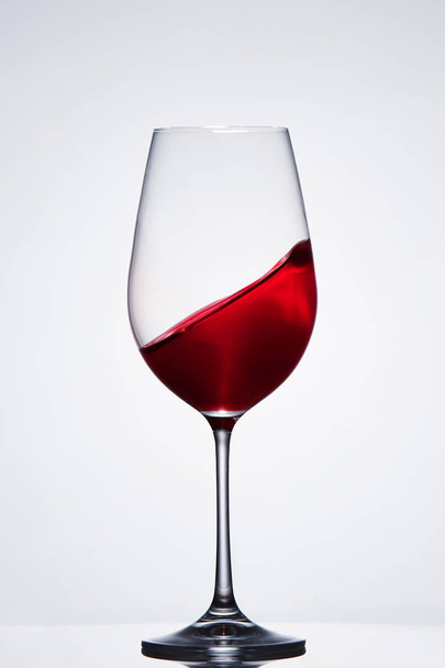 Moving red wine in the pure fragile wineglass standing against light background with reflection. - Zdjęcie, obraz