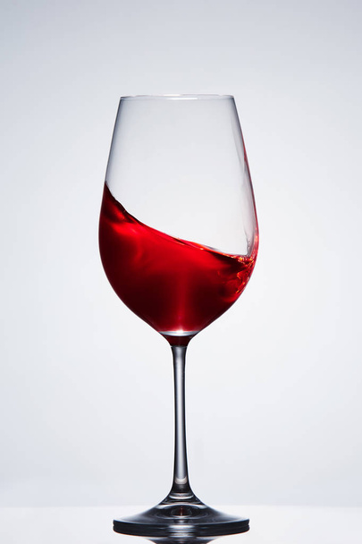 Wave of the red wine in the pure fragile wineglass standing against light background with reflection. - Photo, Image
