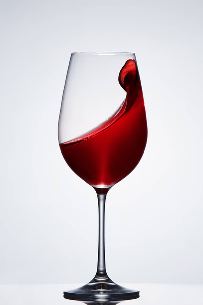 Wave of the romantic drink red wine on the pure wineglass standing against light background with reflection. - Photo, Image