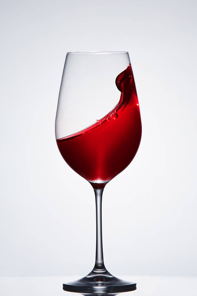 Red wine splash in the delicate elegant wineglass standing against light background. - Photo, Image