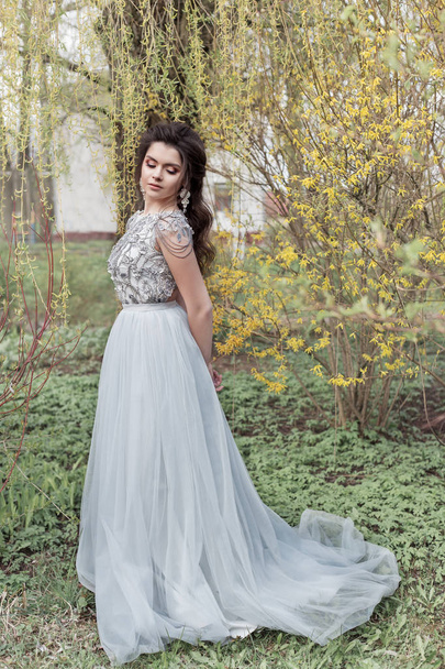 Beautiful young tender gay girl with a beautiful wedding hairstyle with a snow-white smile in a light dress is walking in a spring blooming garden - Foto, imagen