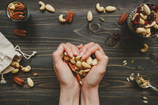 Hands holding nuts in heart shape on the wooden table. Nuts mix with seeds and dried fruits on the wooden table - Foto, Imagem