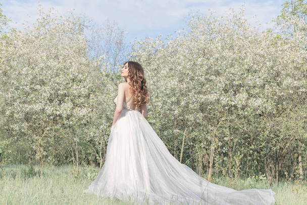 Beautiful sweet girl bride in a tender air wedding dress in a blooming spring garden in the rays of sunlight at sunset. Fine art style - Photo, image