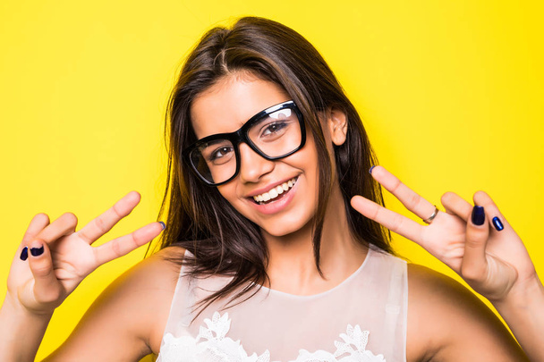 Молода дівчина носить окуляри з жестом перемоги на жовтому тлі. Дівчина дивиться на камеру
. - Фото, зображення