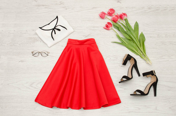 Divat fogalmát. Piros szoknya, blúz, napszemüveg, rúzs, fekete cipő és rózsaszín tulipánok. Felülnézet, világos fa háttér - Fotó, kép