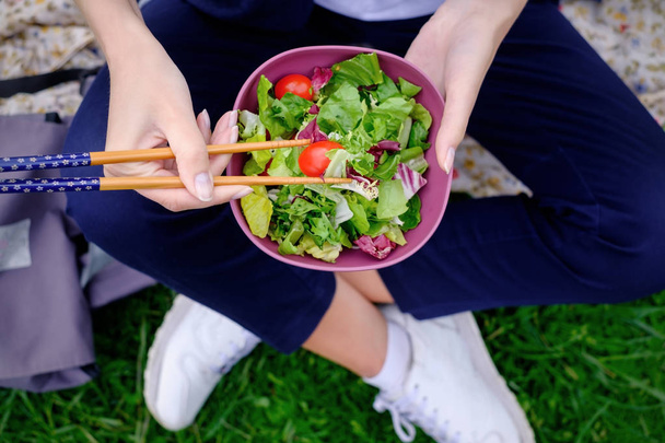 Uma menina senta-se na grama e come uma salada fresca verde com costeletas
 - Foto, Imagem