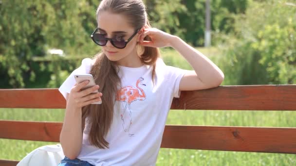 Girl talking on the smart phone sitting in a park on wood bench with green tree leaf, Nature scene - Footage, Video