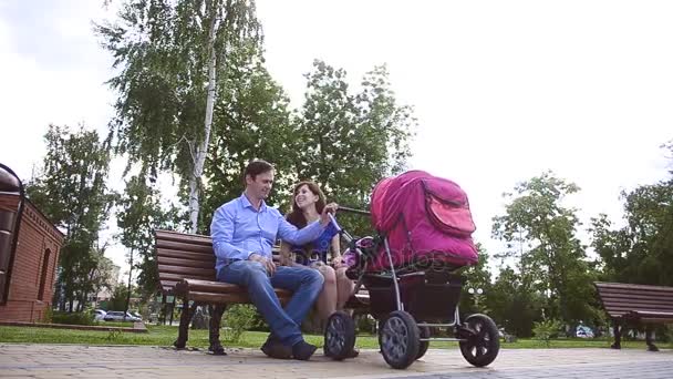 Μαμά και ο μπαμπάς να ξεκουραστούν σε ένα παγκάκι στο πάρκο με καροτσάκι μωρού. - Πλάνα, βίντεο