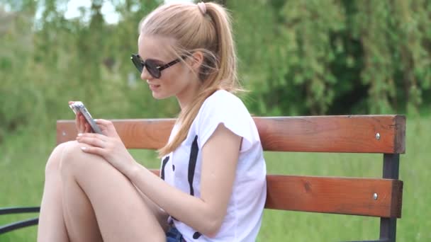 Girl talking on the smart phone sitting in a park on wood bench with green tree leaf, Nature scene - Metraje, vídeo