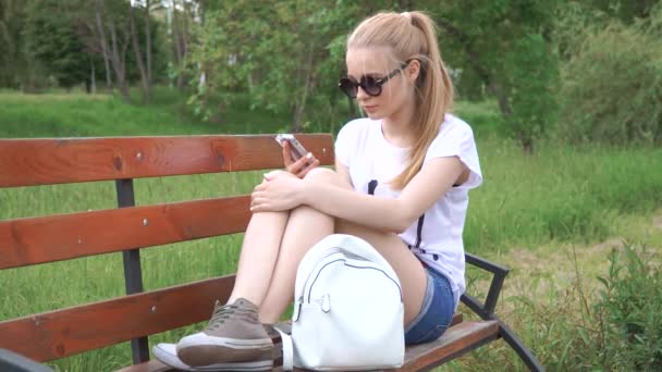 Girl talking on the smart phone sitting in a park on wood bench with green tree leaf, Nature scene - Materiał filmowy, wideo