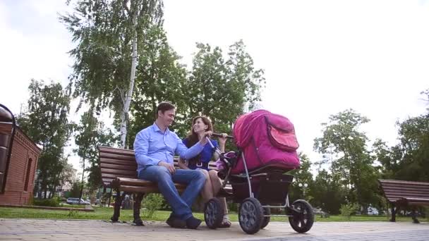Ο μπαμπάς και η μαμά κάθεται σε παγκάκι με καροτσάκι μωρού στο πάρκο, οικογενειακές διακοπές ημέρα το καλοκαίρι. - Πλάνα, βίντεο