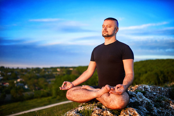 Portrait of the man sitting on a rock in the lotus position against blue sky. - Фото, изображение