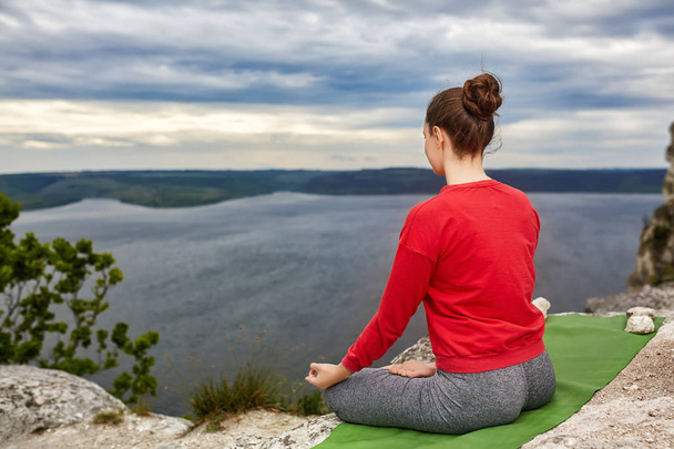 Молодая женщина медитирует в позе лотоса на скале над большой рекой
. - Фото, изображение