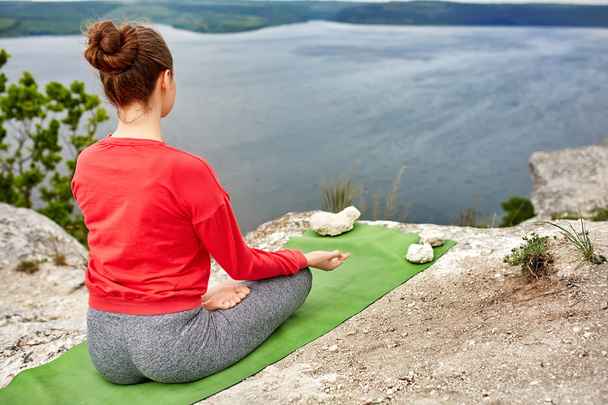 Young woman meditating in a lotus pose on the rock above big river. - Photo, Image