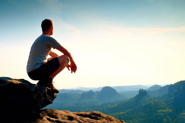 Młody człowiek w czarnym sportowe spodnie i szara koszula jest siedząc na krawędzi urwiska i patrząc na mgliste doliny poniżej - Zdjęcie, obraz