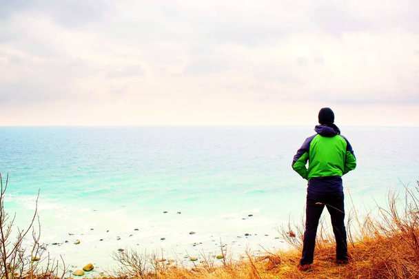 Людина в зеленій вітряній вітрильниці стоїть на скелі над затокою і лоокі на морському далекому горизонті
. - Фото, зображення