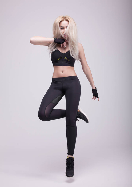 Beautiful young blonde fit woman with long hair, happily jumping on a grey background. Slim woman in sport clothes. - 写真・画像