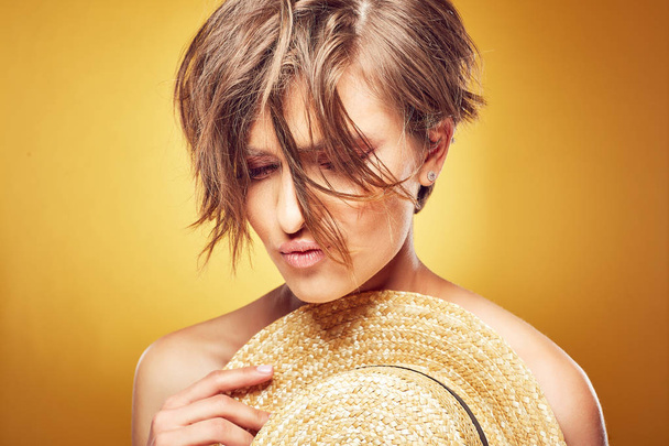 Sexy young pretty woman with short disheveled hair and seductive lips holding straw hat and posing in studio on yellow background - 写真・画像