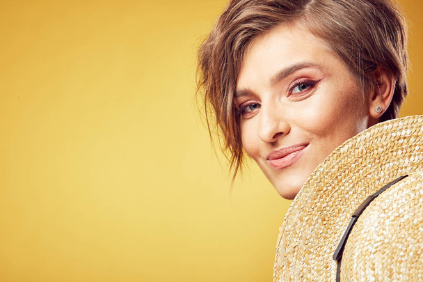 Closeup portrait of sexy young pretty woman with short disheveled brown hair looking at camera with happy face, holding straw hat, posing in studio on yellow background - Zdjęcie, obraz