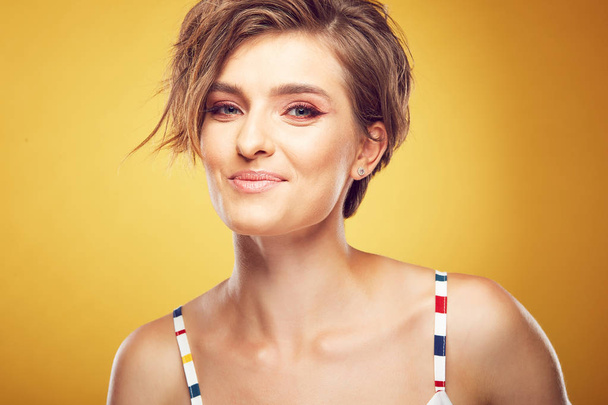 Closeup portrait of sexy young pretty female model with short disheveled brown hair, looking at camera with tender smile, posing in studio on yellow background - Foto, imagen