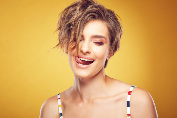 Closeup portrait of sexy happy young pretty female model with short disheveled brown hair, with closed eyes, wide smile and showed tongue, posing in studio on yellow background - Fotó, kép