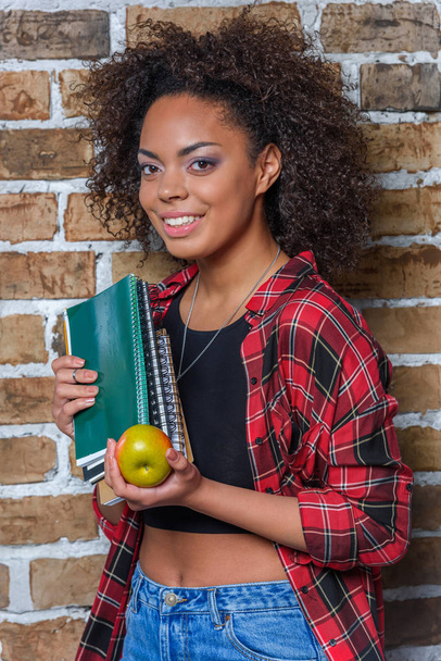 africká americká dívka drží jablko a poznámkové bloky při pohledu na fotoaparát - Fotografie, Obrázek