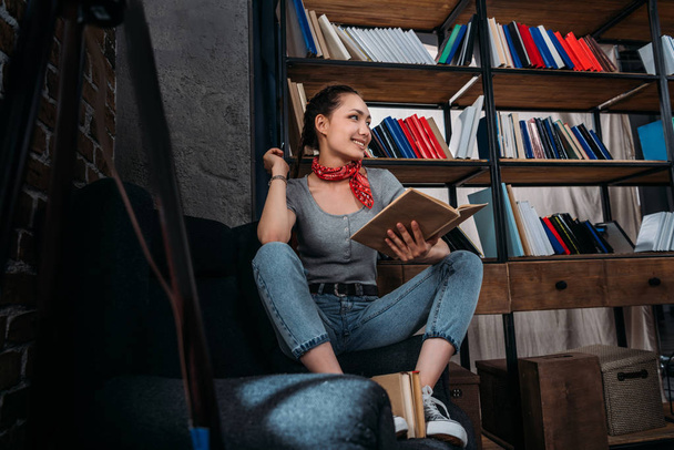 Sorrindo jovem estudante bonita sentada na cadeira com livro e olhando para longe
 - Foto, Imagem