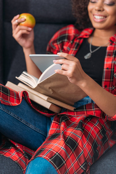 Улыбающаяся молодая женщина сидит в кресле с книгами и с помощью цифрового планшета
 - Фото, изображение
