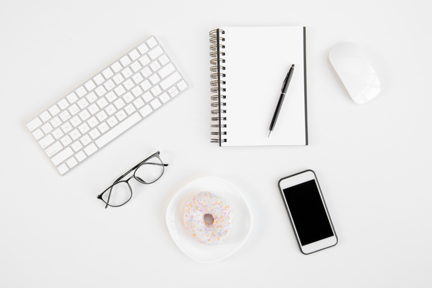 Top view of smartphone with blank screen, blank notebook with pen, eyeglasses, keyboard, computer mouse and tasty doughnut at workplace - Фото, изображение