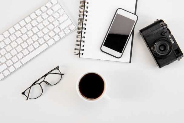 Top view of cup of coffee, keyboard, notebook, eyeglasses, smartphone and camera at workplace - Fotó, kép