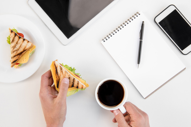 Top view of person eating sandwiches and drinking coffee at workplace with electronics  - Photo, Image