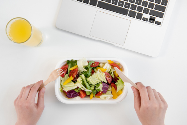 Cropped shot of person eating fresh salad and drinking orange juice at workplace with laptop - Photo, Image