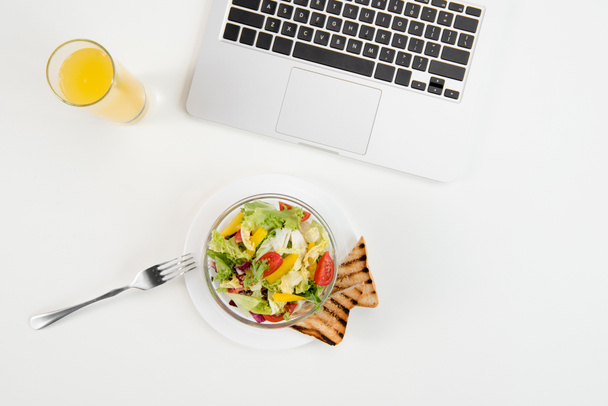 Top view of laptop, orange juice in glass and fresh healthy salad with toasts at workplace - Photo, Image