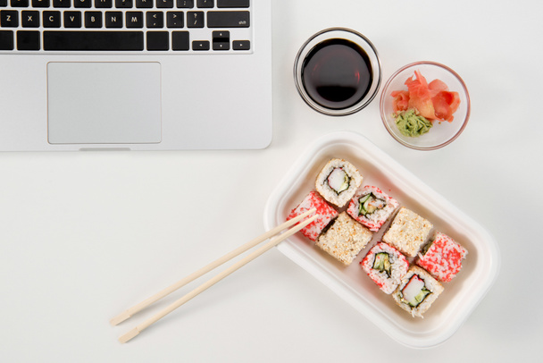 Bovenaanzicht van een opengeklapte laptop en sushi rolt met wasabi en sojasaus op werkplek - Foto, afbeelding
