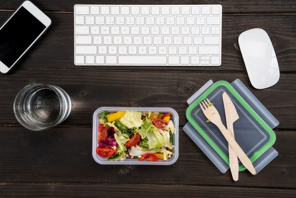 Ansicht von Smartphone, Computertastatur und Computermaus mit frischem Salat auf Holztisch.  - Foto, Bild