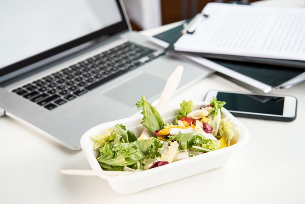 close up of fresh salad with laptop and smartphone on tabletop at workplace - Fotó, kép