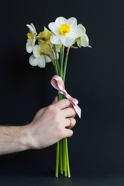 Precioso ramo de narciso blanco y amarillo con un lazo hecho de cinta rosa en la mano de un hombre casado sobre un fondo negro. Agradable para postal
 - Foto, Imagen