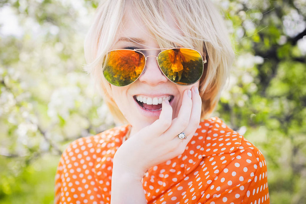 portrait outdoors young beautiful girl in an orange hipster blonde bright cheerful  dot blouse and smiling in  sunglasses on a background blooming trees with white teeth - Foto, immagini