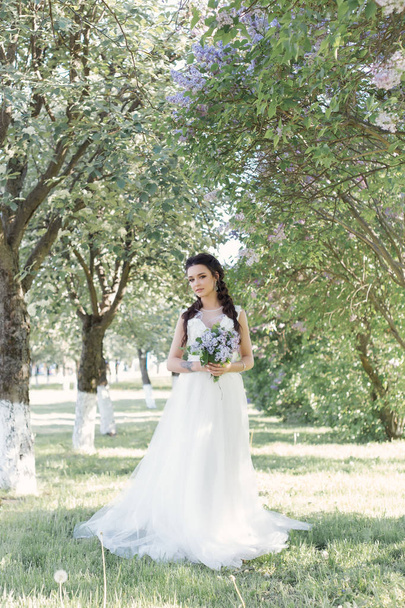 Tender beautiful cute girl bride in a white air dress with a bouquet of lilacs in her hands walking through the park on a sunny spring day. Photo in gentle colors - 写真・画像